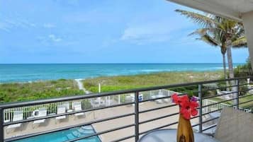 A balcony with a view of the ocean, a pool, and a table with a vase of flowers.