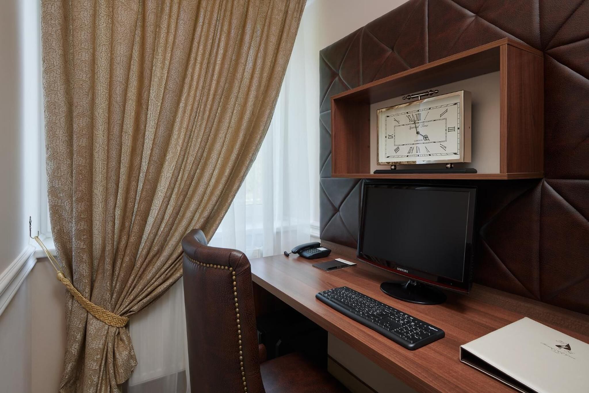 A hotel room with a wooden desk, a computer, a telephone, and a wall-mounted clock.