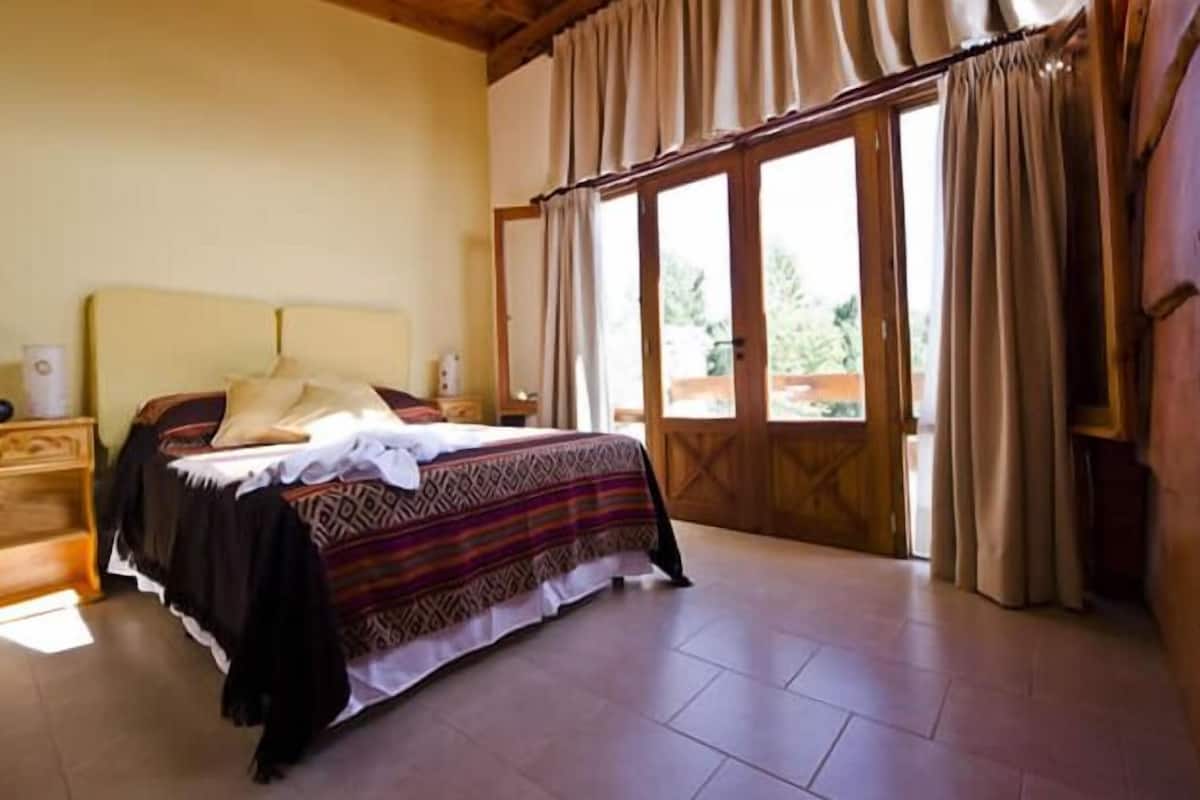 Un dormitorio con cama, mesita de noche, ventana con cortinas y vistas al exterior.