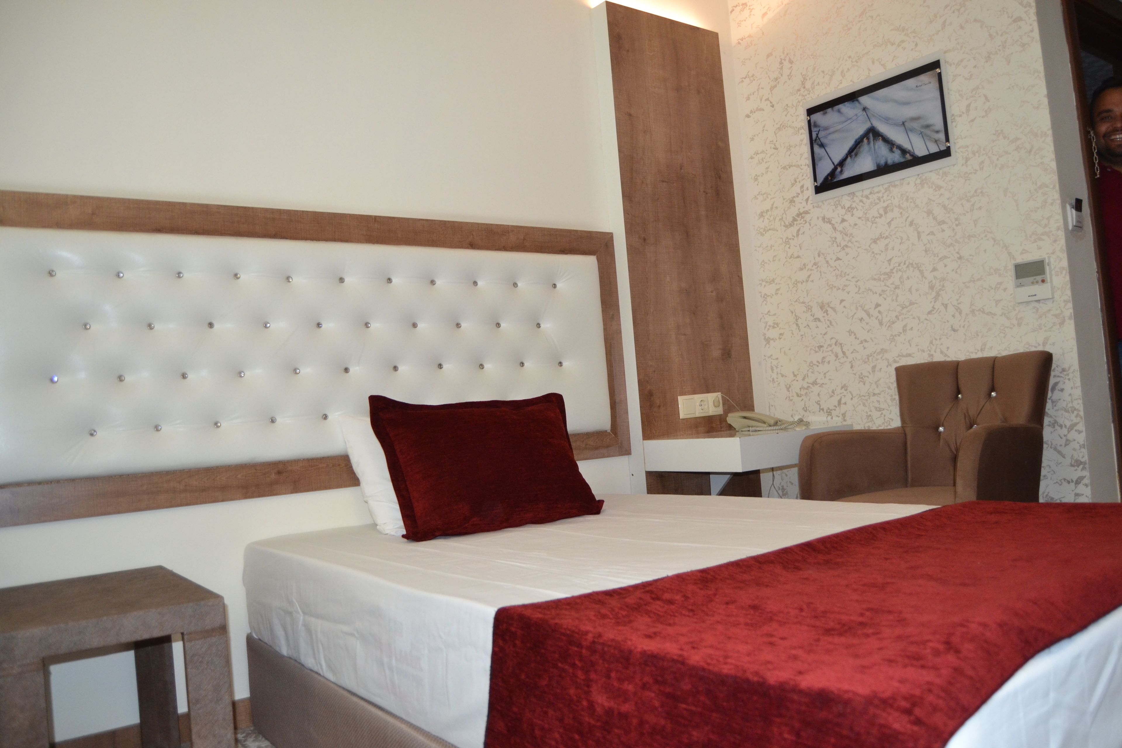 A hotel room with a tufted headboard, a red throw blanket, a wooden nightstand, and a desk with a chair.