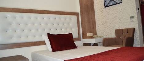 A hotel room with a tufted headboard, a red throw blanket, a wooden nightstand, and a desk with a chair.
