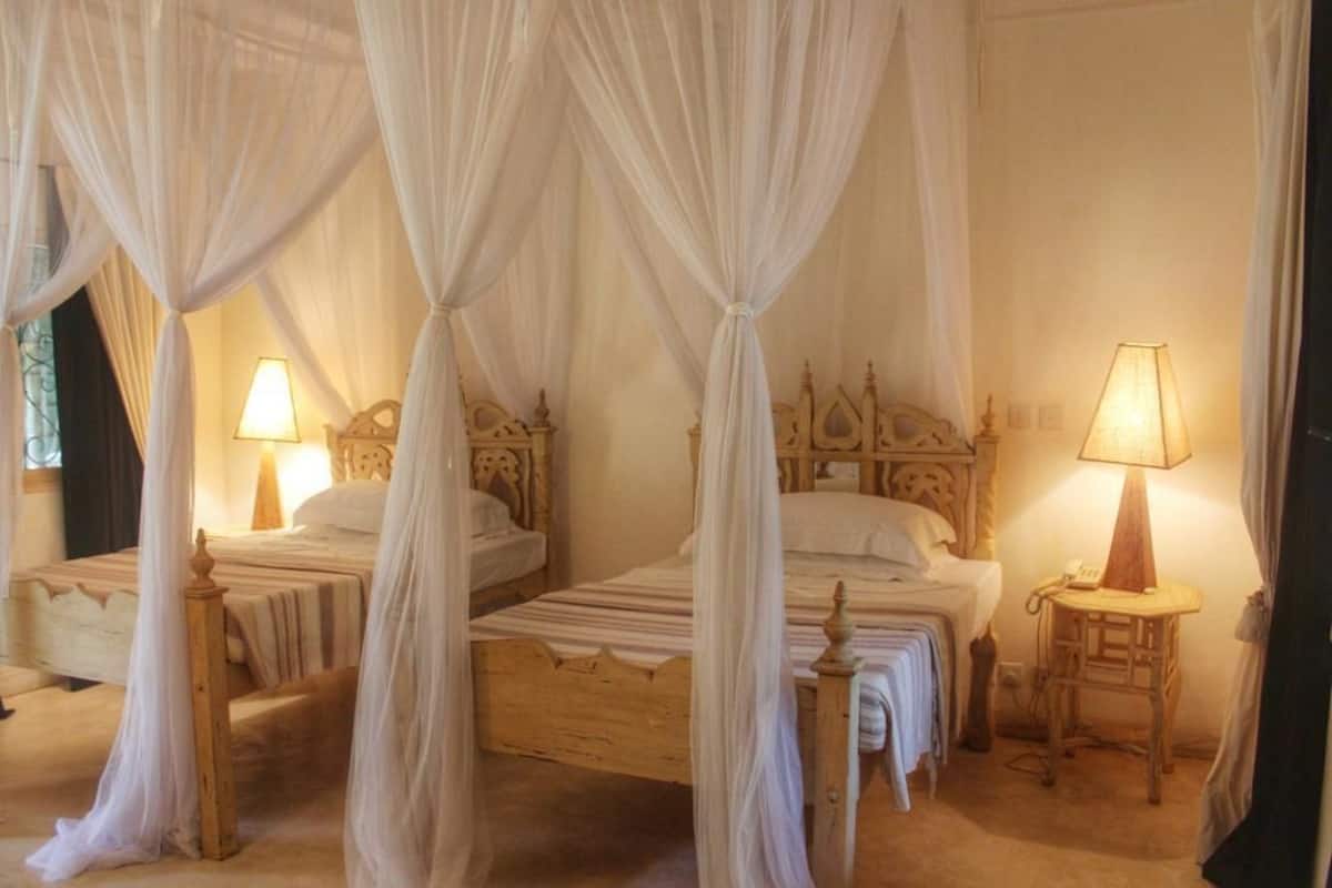 Ein Zimmer mit zwei hölzernen Betten, jedes mit Baldachin. An den Betten stehen Nachttische mit Lampen.