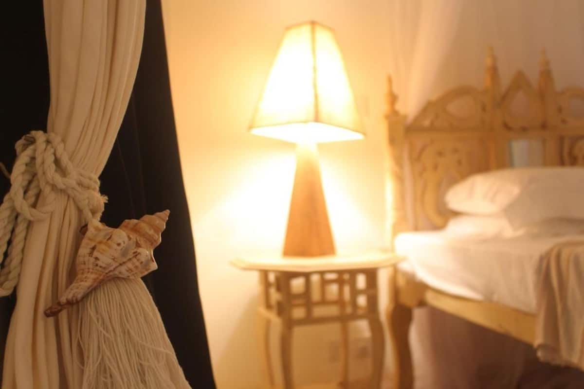 Ein Bett mit Holz-Kopfteil, ein Nachttisch mit Lampe und ein Vorhang mit Kordel- und Muscheldekoration.