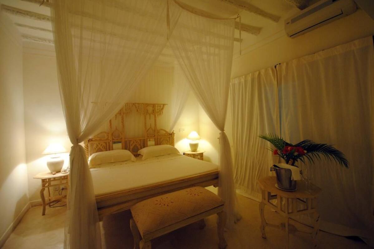 Ein Schlafzimmer mit Himmelbett, Nachttischlampen, einem kleinen Tisch mit einer Vase und einer Bank.