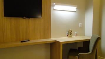 A room with a wooden desk, a chair, a television mounted on the wall, and a bedside table with a lamp and a box.