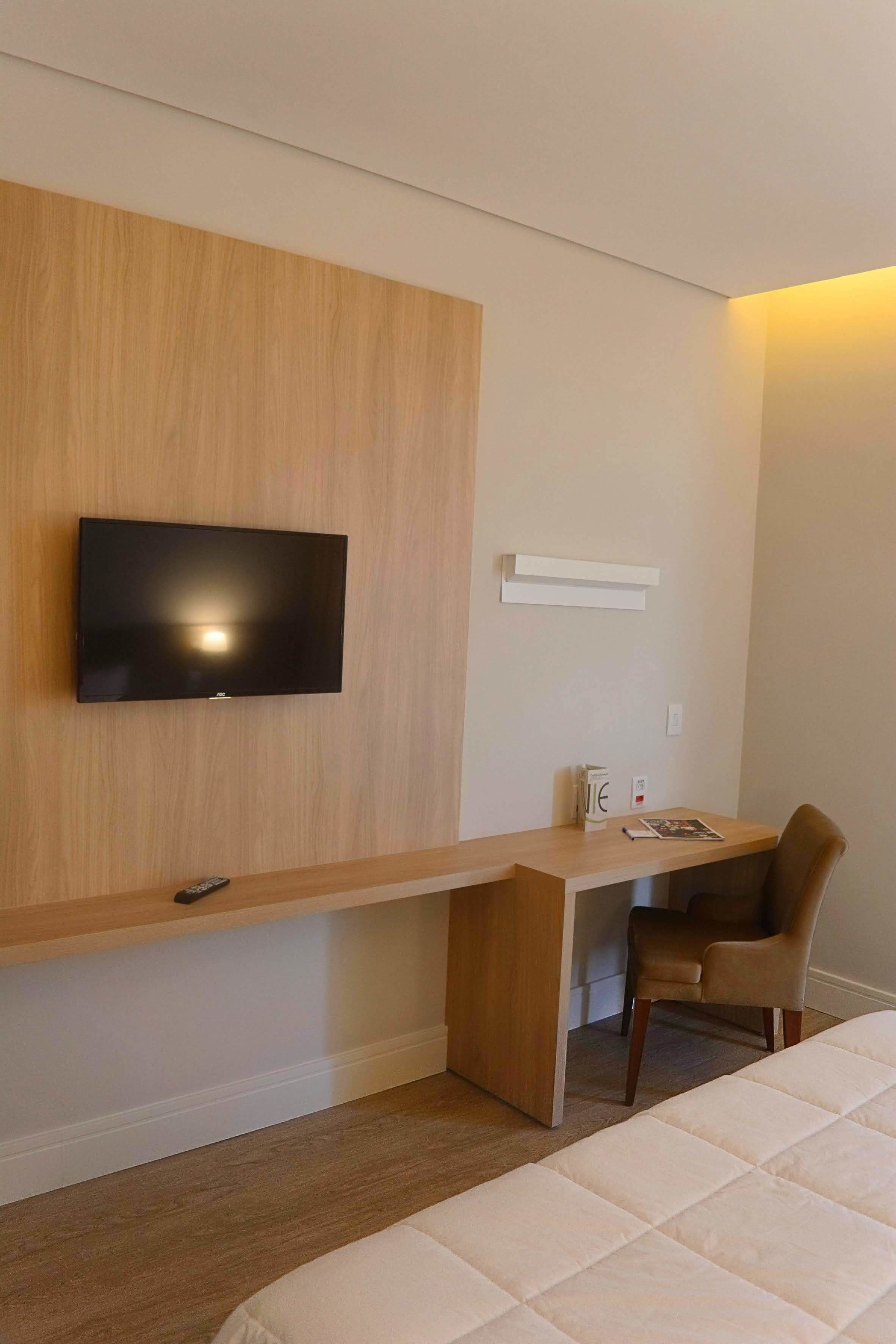 A hotel room with a wooden desk, a television mounted on the wall, a chair, and a bed.