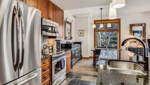 A modern kitchen with stainless steel appliances, wooden cabinets, and a large window offering a view of trees. - Capitol Peak Lodge 3313 (Snowmass Village)
