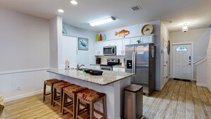 A modern kitchen with white cabinets, a stainless steel refrigerator, a microwave, and a sink. There are three wooden bar stools with red and beige upholstery. - Shared Heated Pool, Garage Access, Golf Cart Included, Pet Friendly (Port Aransas)