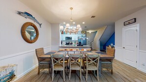 A dining area with a wooden table and chairs, a chandelier, and a kitchen in the background. - Shared Heated Pool, Garage Access, Golf Cart Included, Pet Friendly (Port Aransas)