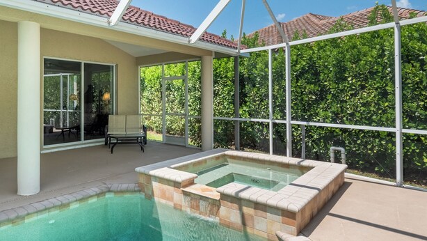 A swimming pool with a tiled deck, a patio area with a table and chairs, and a view of green foliage through large windows.