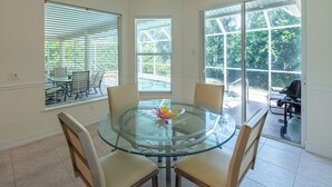 A glass-top dining table with four chairs, large windows with blinds, and a view of a patio area.