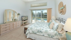 Un dormitorio con cama, televisor, cómoda, ventana con vista a una piscina y follaje, y ventilador de techo.