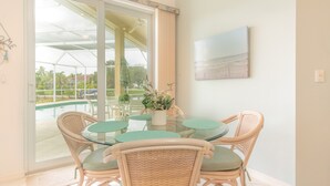 Een eethoek met een glazen tafel, rieten stoelen, een kamerplant en uitzicht op een zwembad en groen buiten.