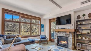 A modern living room with a stone fireplace, large windows, a flat-screen TV, and a cozy seating area. - Capitol Peak Lodge 3417 (Snowmass Village)