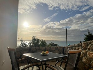 A table with chairs on a balcony overlooking a body of water, with a stone wall and vegetation in the background. - Apartments Natasa (Starigrad)