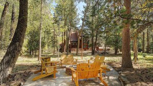 Una terraza de madera con sillas Adirondack y un hogar de piedra en un área boscosa.