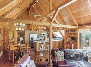 A spacious cabin interior with a wooden ceiling, a dining area, a living room with a leather sofa, and a kitchen area.
