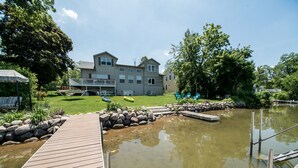 A wooden dock leading to a calm pond with a stone border, surrounded by lush greenery and a large building with a balcony in the background. - Large Lakefront Home on Lake Como - Private Pier! (Lake Geneva)