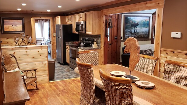 A cozy cabin interior with wooden walls, a dining area with wicker furniture, and a kitchen with modern appliances. - Beautiful Home in the Woods on Bike Trail (Lake George)