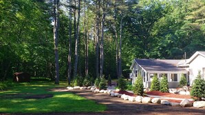A house surrounded by a forest with a well-maintained lawn and a stone pathway leading to the entrance. - Beautiful Home in the Woods on Bike Trail (Lake George)