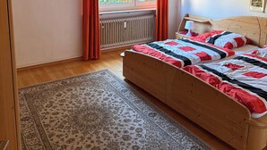 Une chambre avec un lit en bois, une grande fenêtre avec des rideaux, un radiateur et un tapis.