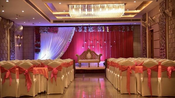A banquet hall with rows of chairs covered in white and red sashes, a stage with a red curtain and floral decorations, and a chandelier. - Orchid Blu (Aligarh)