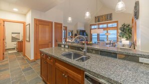 A spacious kitchen with granite countertops, a double sink, wooden cabinets, pendant lighting, and a view through large windows.