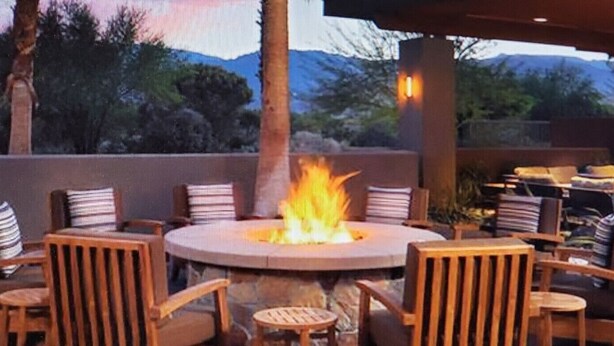 A round table with chairs around a central fire pit, set in an outdoor patio area with a view of trees and mountains in the background.