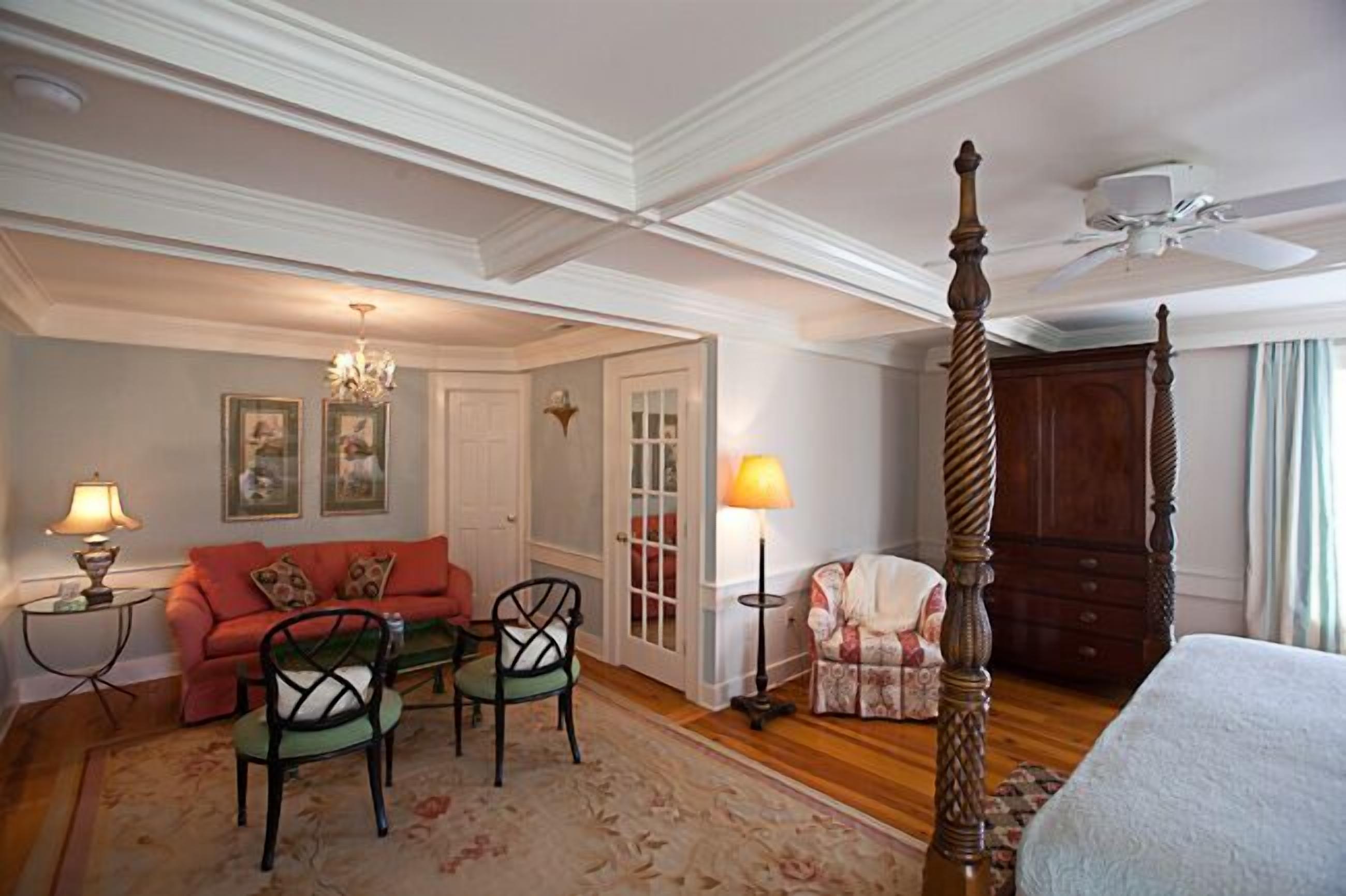 A bedroom with a four-poster bed, a sitting area with a sofa and chairs, a chandelier, and a door leading to another room.