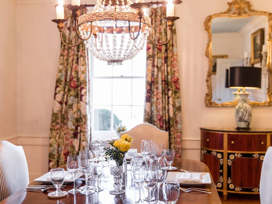 Una mesa de comedor dispuesta para una comida, con una araña, cortinas florales y un espejo en la pared.