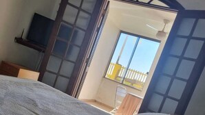 A bedroom with a bed, a TV on a stand, and a window with a view of the outside. - Pousada Girassol (Rio das Ostras)