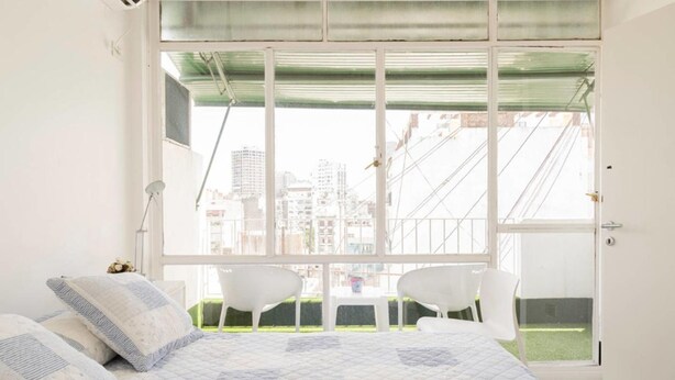 A bedroom with a large window, a bed with a quilt, two white chairs, and a small table. - Luminoso Duplex en Recoleta (Buenos Aires)