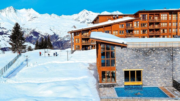 A hotel with a pool, ski slope, and snow-covered mountains in the background.
