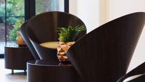 A modern interior with a dark-colored chair with a unique curved design, a small potted plant on the chair, and a view of greenery outside through large windows. - Alife Apart Hotel (Beirut)