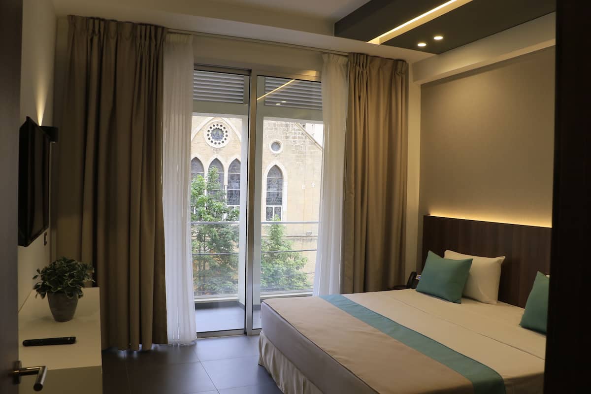 Une chambre d’hôtel avec un grand lit, une télévision, une plante en pot et une vue sur un immeuble aux fenêtres en arc de cercle.