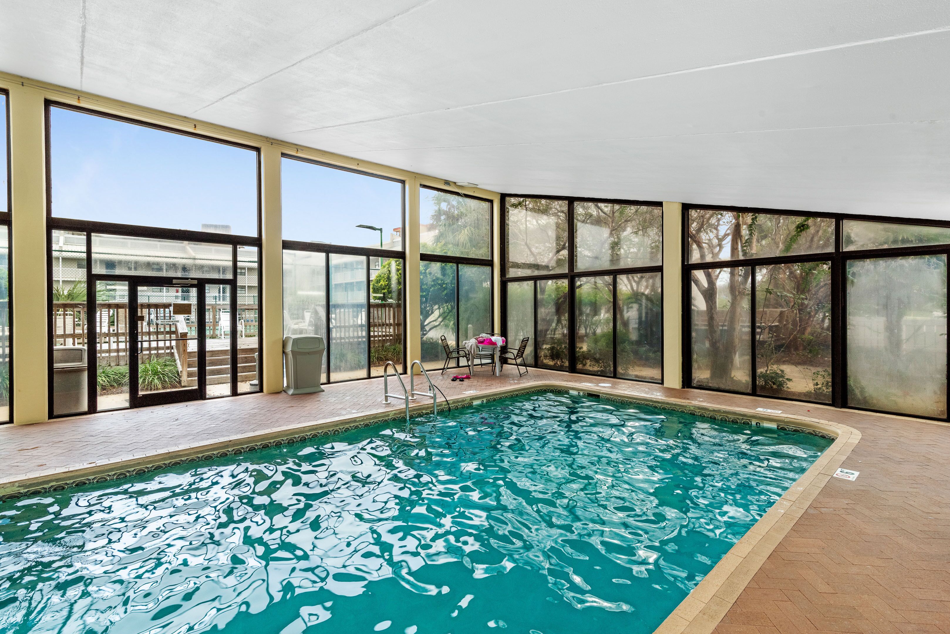Una piscina interna con pareti di vetro trasparente, pavimento piastrellato e alcuni sedili e tavoli nella zona della piscina.