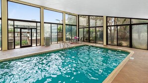An indoor swimming pool with clear glass walls, a tiled floor, and a few chairs and tables around the pool area.