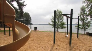 Un parque infantil con tobogán y barras colgantes, cerca de un cuerpo de agua y árboles al fondo.