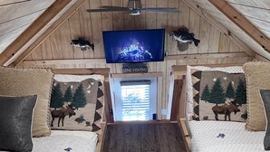 Una habitación cálida tipo cabaña con dos camas, un ventilador de techo, un televisor y una ventana con persianas.