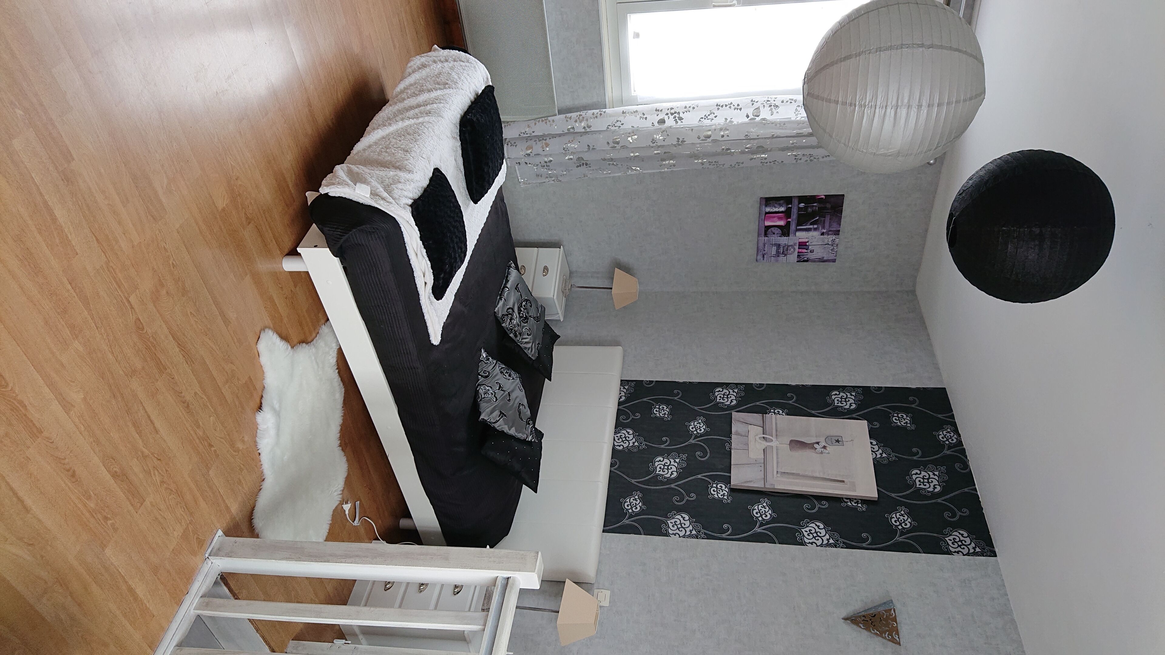 Een kamer met een houten vloer, een witte bank, een tapijt met een patroon, een kleine tafel met een lamp en een raam met uitzicht.