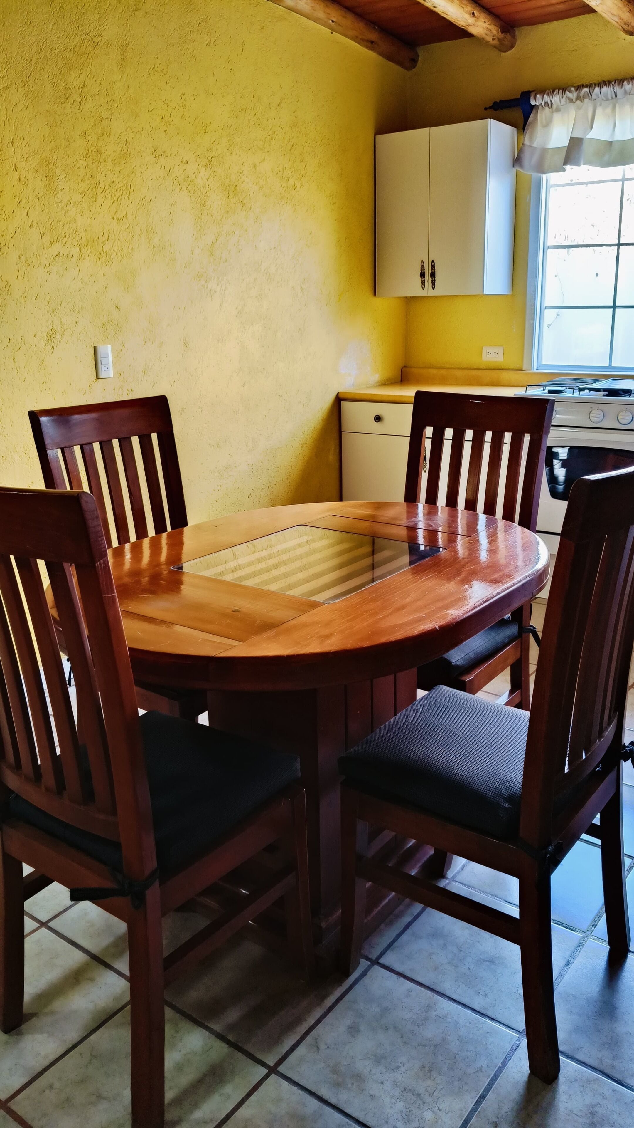 A dining area with a wooden table and chairs, a kitchen in the background, and a window with curtains.