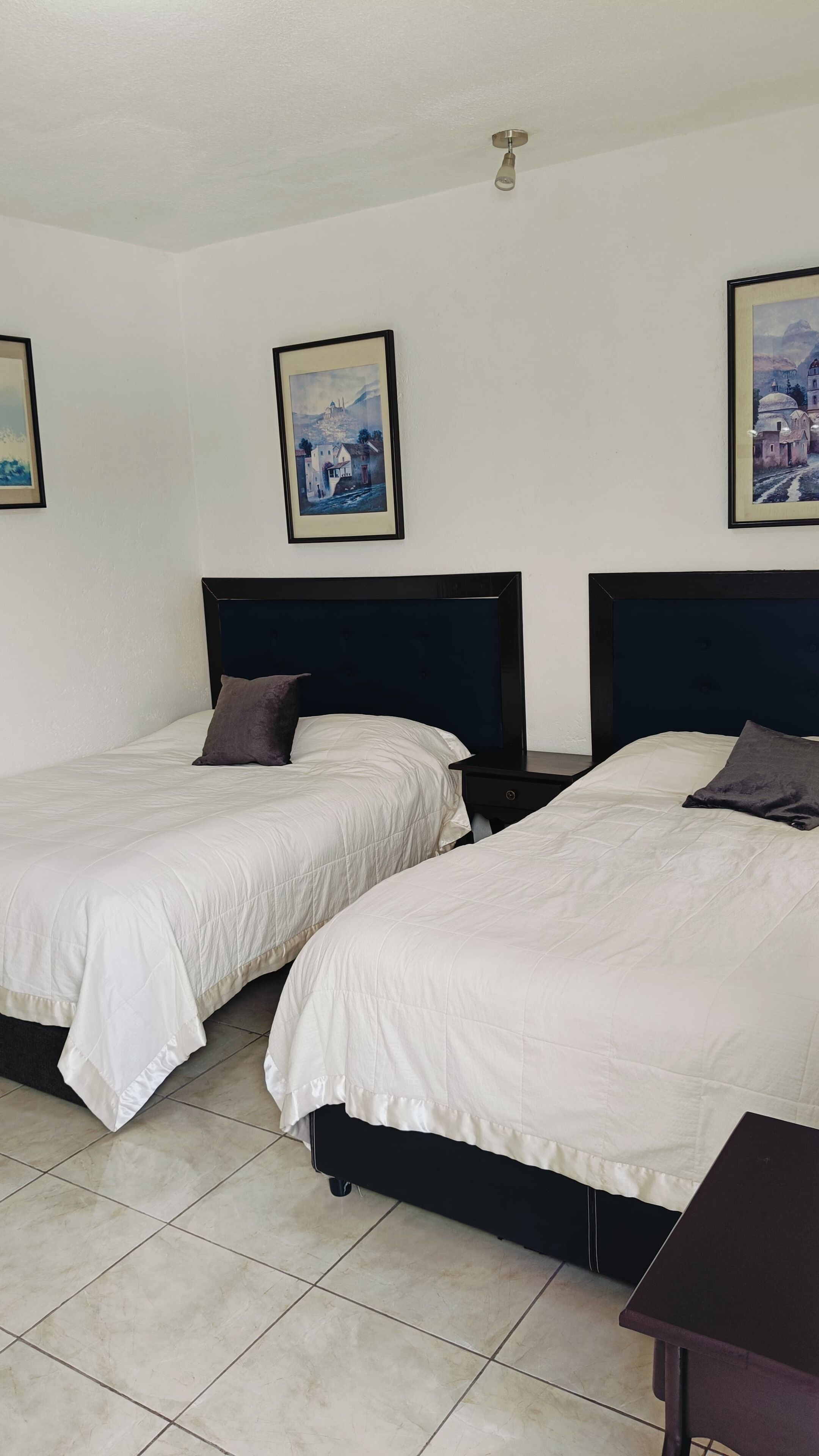 Camera d'albergo con due letti, un comodino e un tavolino. Alle pareti, si vedono quadri incorniciati.