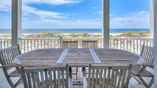 Une terrasse en bois avec une table et des chaises, donnant sur une plage bordée de dunes et l’océan.