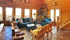 A log cabin interior with a wooden staircase, a blue leather sofa, a wooden rocking chair, and a stone fireplace. - Red Rock Ranch Log Cabin: Large, Fully Furnished, 5 Bdr, sleeps 12, 3 levels (Escalante)