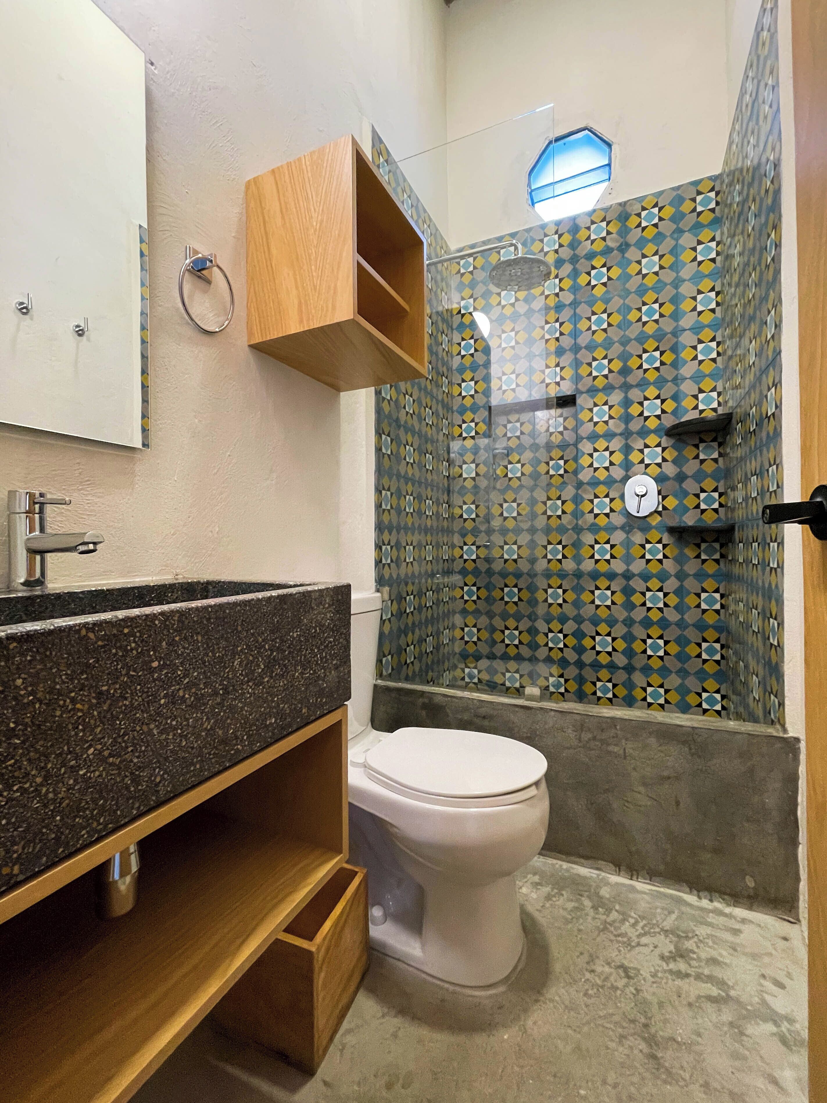 Un baño con encimera de granito, inodoro blanco, mueble de madera y ducha con azulejos estampados.