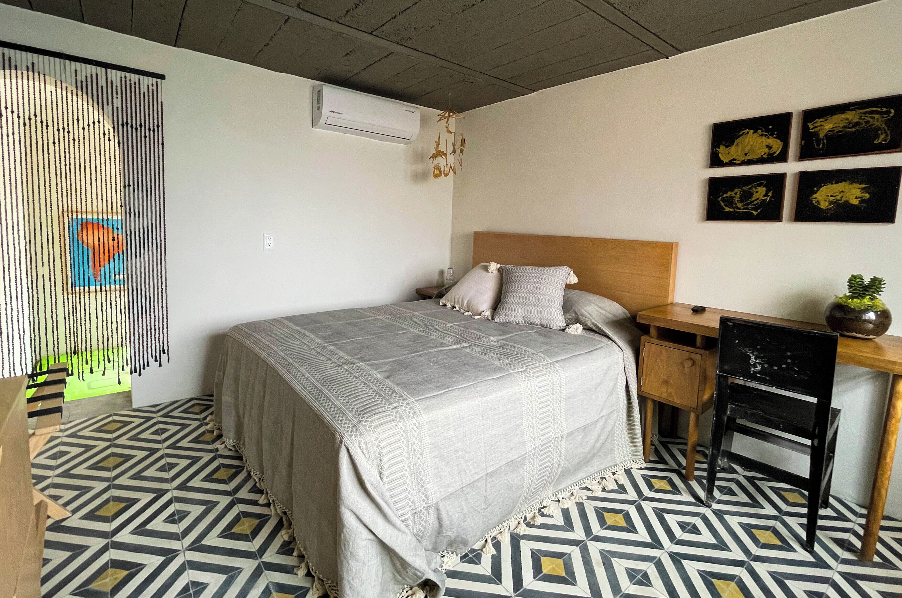 A bedroom with a bed, a desk, and a chair. There is a wall-mounted air conditioner and a wall art piece.