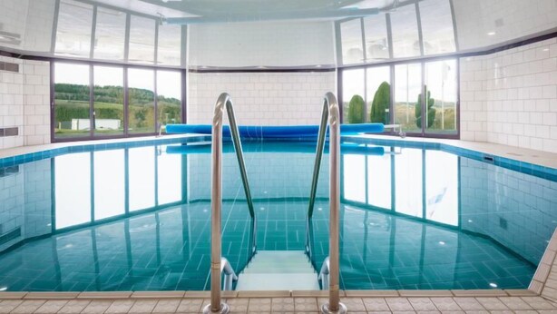 An indoor swimming pool with clear glass walls, a blue tiled floor, and metal handrails. - Holzchalet A-D by Interhome (Gerolstein)