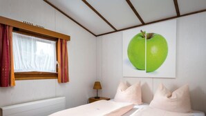 A bedroom with two beds, a window with curtains, a lamp, and a wall-mounted apple artwork. - Holzchalet A-D by Interhome (Gerolstein)