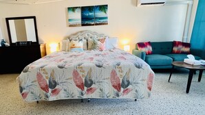 A hotel room with a large bed, a sofa, a small table, a dresser, a mirror, and a painting of a beach scene. - Condo with King Size Bed, near Downtown, with Ocean Views, AC, and Secured. (Christiansted)