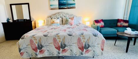 Une chambre d’hôtel avec un grand lit, un canapé, une petite table, une commode, un miroir et un tableau représentant une scène de plage.
